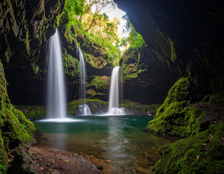 Dual showing dual waterfalls flowing into a dark green pool from a cave entrance with mossy rocks keywords:...の素材