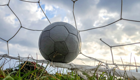 A gray soccer ball rests in a net with a cloudy sky and green grass in the background.の素材