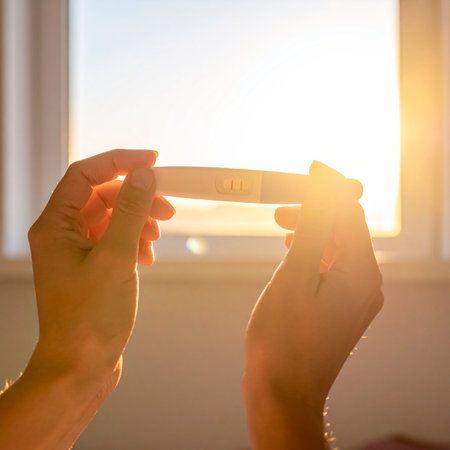 Close-up of hands holding a pregnancy test showing two lines, illuminated by bright window light.の素材