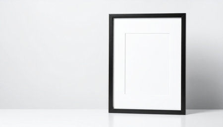 A black framed picture frame with a white mat and blank white space sits on a white surface with a light gray background.の素材