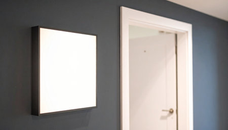 A blank white square sign is mounted on a dark gray wall next to a white interior door in a softly lit hallway.の素材