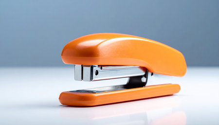 A bright orange stapler is placed on a white surface against a soft blue gradient background.の素材