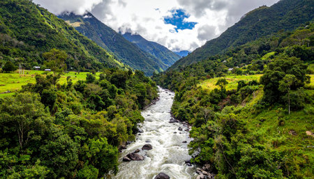 An expansive river valley carved between steep, green mountains, with a winding river flowing through lush vegetation under a cloudy sky.の素材