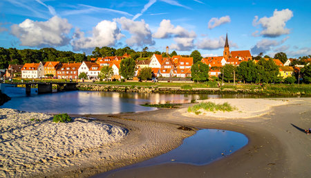 A sandy beach meets a calm waterway, with a European town featuring a prominent church steeple and red-roofed houses visible across the water.の素材