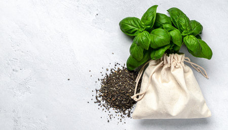 A small fabric drawstring bag filled with dried loose leaf tea, with a pile of spilled tea and fresh basil leaves beside it on a light gray surface.の素材