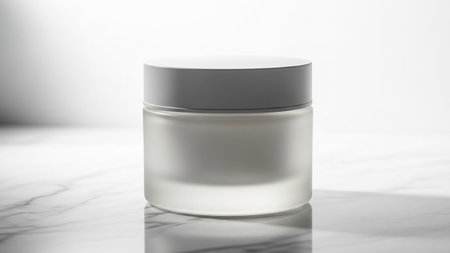 A frosted glass cosmetic jar with a white lid rests on a marble surface, bathed in soft light and subtle shadows.の素材