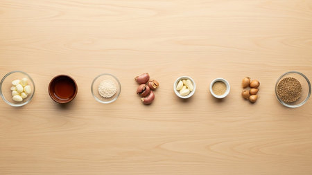 Various cooking ingredients including garlic, shallots, spices, and oil are neatly arranged in small bowls on a light wooden surface.の素材