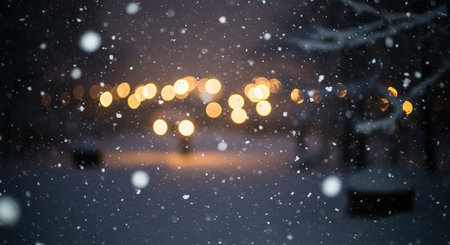 Soft snowflakes fall against a dark background with blurred warm lights creating bokeh.の素材