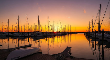 A picturesque marina filled with numerous sailboats silhouetted against a vibrant orange and yellow sunset sky, with their reflections shimmering on...の素材