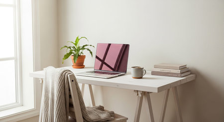 A cozy home office desk with a laptop, plant, books, and a draped blanket near a window.の素材