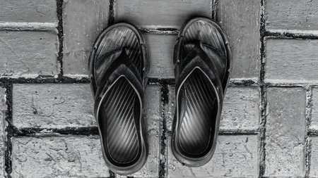 Low-angle view of black rubber flip-flops on textured paving blocks. High-contrast monochrome photography symbolizing simplicity, or a pause in a journey.の写真素材