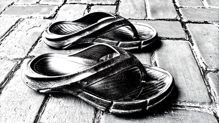 Low-angle view of black rubber flip-flops on textured paving blocks. High-contrast monochrome photography symbolizing simplicity, or a pause in a journey.の写真素材