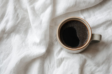 A serene flat lay of a cozy coffee cup filled with rich black brew resting on soft white fabric. Perfect for conveying relaxation and comfort at home.の素材