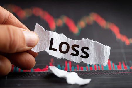 A close-up image of a hand holding a torn piece of paper with the word "LOSS" against a blurred financial chart, symbolizing financial decline and market uncertainty.の素材