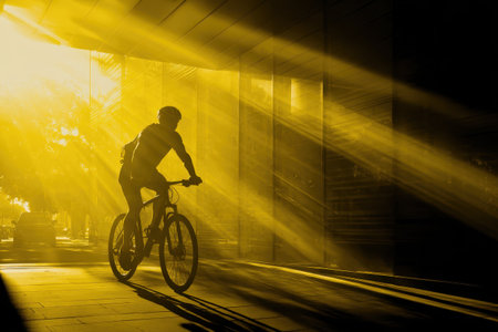 A dramatic silhouette of a cyclist riding through golden sunlight rays in an urban environment. The image captures motion, vitality, and the beauty of outdoor exercise.の素材