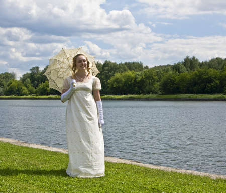 Woman in white historical dress with white umbrella near lake.