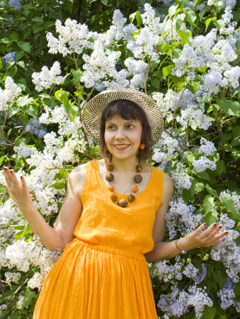 Pretty lady in orange dress in hat brunette European standing near lilac shrub in blossomの写真素材
