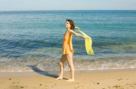 Young European woman with brown hair in orange shirt with yellow scarf on sea beach.の写真素材
