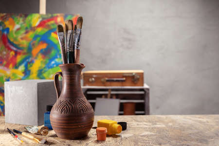 Artist supplies at wooden table. Paint brush in clay jug and art painter tool on desk background texture. Paintbrush for painting in artist workplaceの写真素材