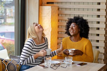 Two multi ethnic girl friends enjoying coffee together in a coffee shop, sitting at the table chatting and laughing out loud.の写真素材