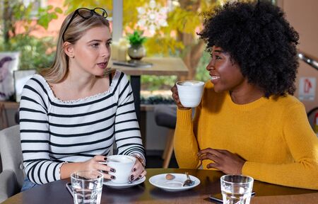 Two young girl friends enjoying coffee together in a coffee shop, sitting at a table, chatting and gossiping. Diversity and multi ethnic concept. No to racism.の写真素材