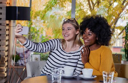 Two young women enjoying coffee together in a coffee shop, sitting at table taking a selfie with a smartphone. Diversity and multi ethnic concept. No to racism.の写真素材
