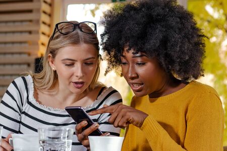 Two young girl friends enjoying coffee together in a coffee shop, sitting at a table, chatting and...