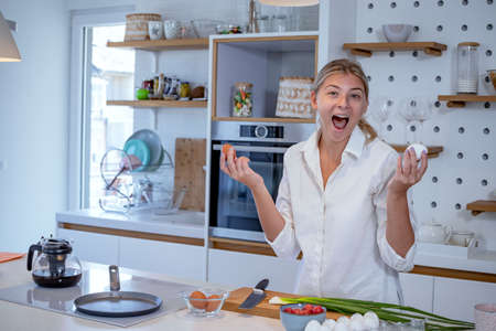 Beautiful young woman cooking in a modern kitchen. Preparing breakfast, holding eggs and choosing between white or a yellow shell egg. Smiling at camera.の写真素材