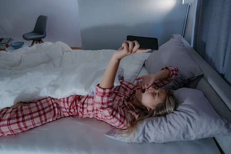 Beautiful young woman in pajamas stretching in bed, turning off the alarm clock on a mobile phone, having a hard time to wake up.の写真素材