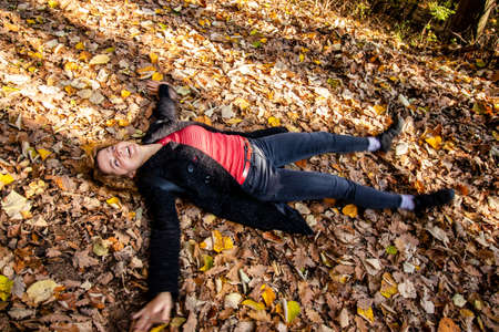 Beautiful laughing woman, lying on a carpet of fallen leaves and making "snow angel" in a lush autumn foliage in the forest. Relaxing and having fun in the woods.の写真素材