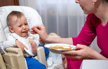 Baby boy sitting in a feeding chair, eating his first food from the spoon with mom at home.