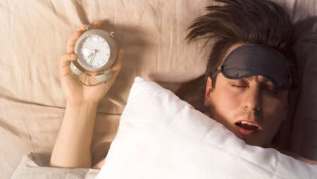 Hands of a young man from under the blankets hold an alarm clock in gray.の写真素材