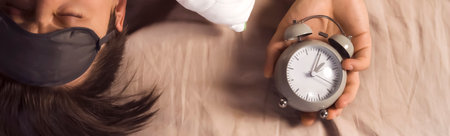 Hands of a young man from under the blankets hold an alarm clock in gray.