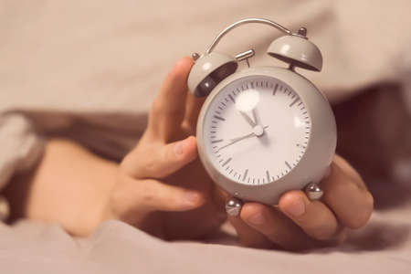 Hands of a young man from under the blankets hold a retro vintage alarm clock.の写真素材