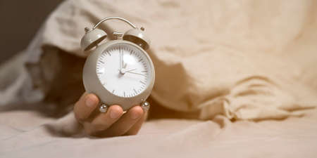 Hands of a young man from under the blankets hold an alarm clock in gray.の写真素材