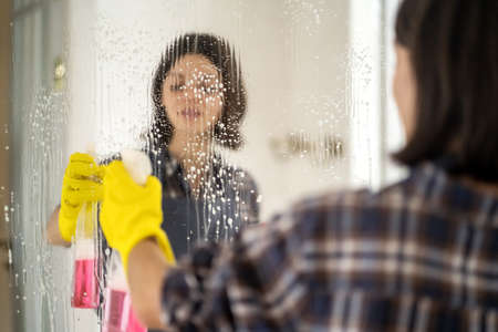 Young woman is washing mirror in the bathrom.
