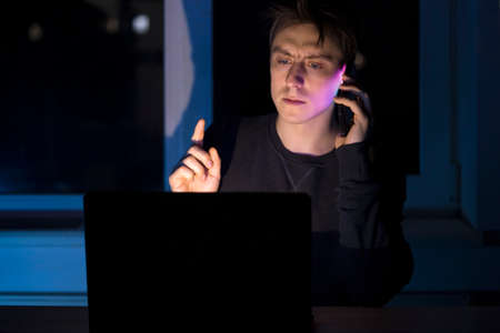 Man works on a laptop at night, talks on phone.