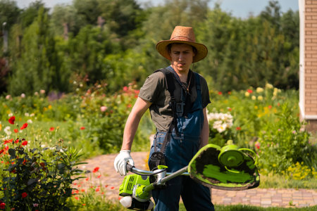 A young white man in a straw hat is mowing a lawn with a lawn mower in his backyard garden