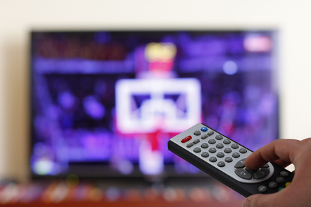 Watching a basketball match in the television, with a tv remote control in the hand