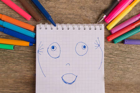 Open white notepad with colorful felt-tip pens and ball pens with drawing glad face on the wooden table.の写真素材