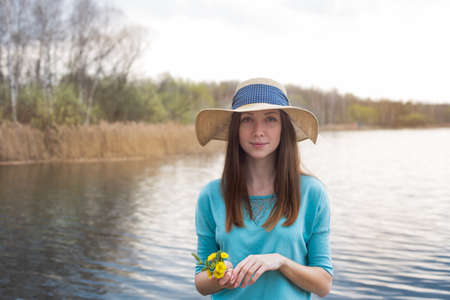 Freckled girl in hat and with bouquet standing on shore of the lakeの写真素材