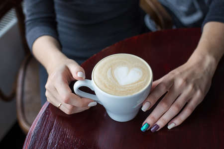 Women's hands holding a cup of coffee with foam and heartの写真素材