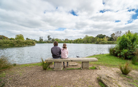 Brother and Sister Sitting on Park Bench in Front of a Lake in Auckland New Zealand - with copy spaceの写真素材