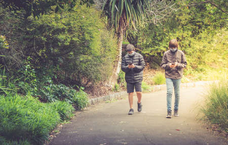 Teenage Mixed Race Brothers Walking on Path In Park Using Their Mobile Phones - with copy space