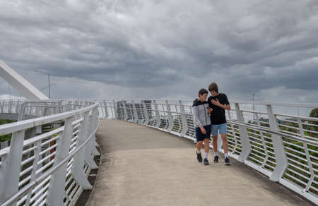Wide Shot of Two Teenage Boys Looking at a Mobile Phone While Walking Over a Bridge in a City with Copy Spaceの写真素材