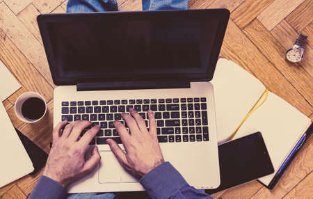 Man working on a wooden floor with his laptop, phone, tablet and agenda.