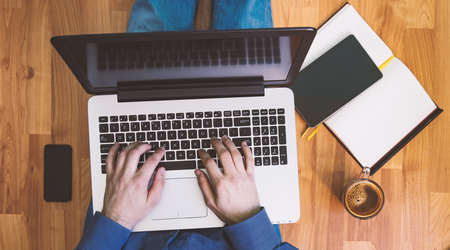 Man working on a wooden floor with his laptop, phone, tablet and agenda.