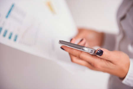 Business woman holding a smart phone and a business report in background - business concept.の写真素材