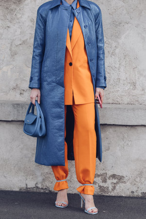 Milan, Italy - February, 24: Street style, woman wearing a blue shiny leather long coat, a matching blue handbag, a neon orange blazer jacket, matching neon orange suit pants and pale blue heels sandals.のeditorial素材