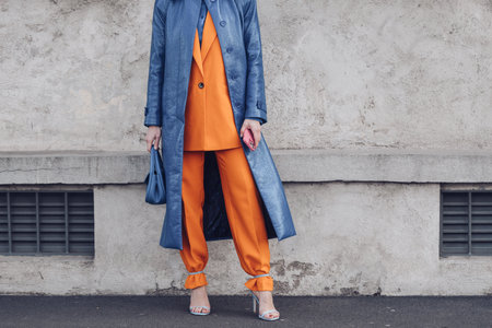 Milan, Italy - February, 24: Street style, woman wearing a blue shiny leather long coat, a matching blue handbag, a neon orange blazer jacket, matching neon orange suit pants and pale blue heels sandals.のeditorial素材